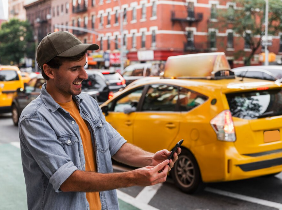 smiling man holds a smartphone on the city street, the yellow cabs on the city road and orange building behind him 