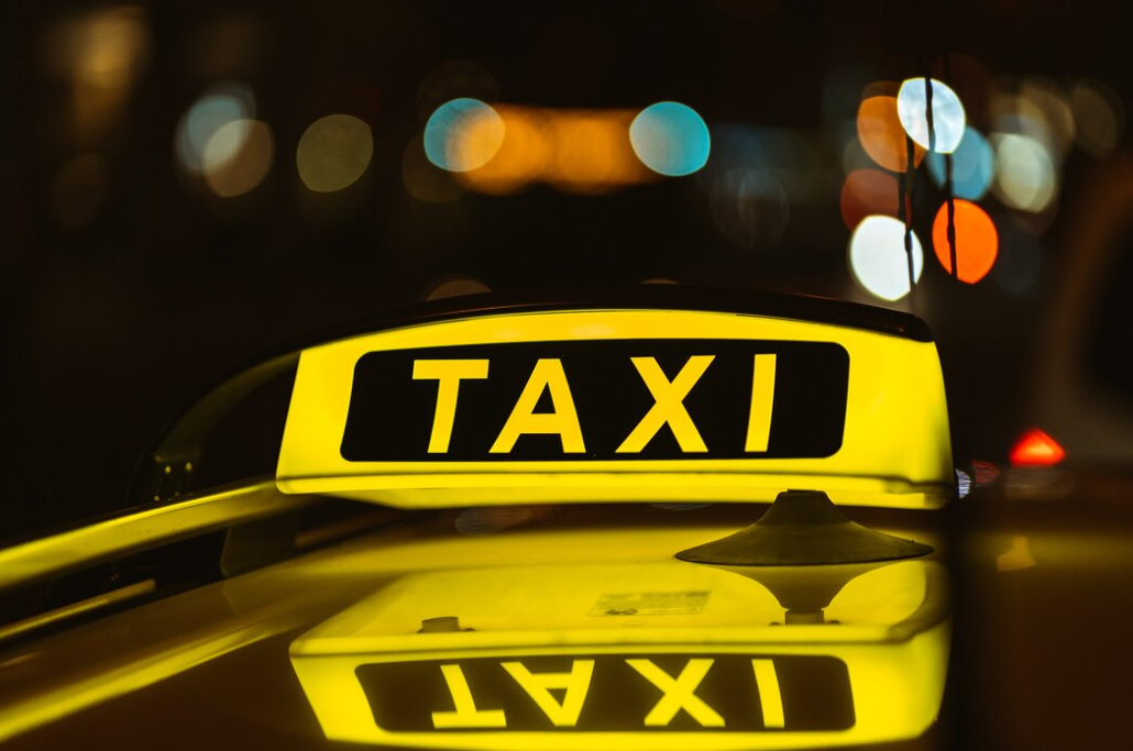 Black and yellow sign of taxi at night placed on top of a car, city lights behind on blurred background